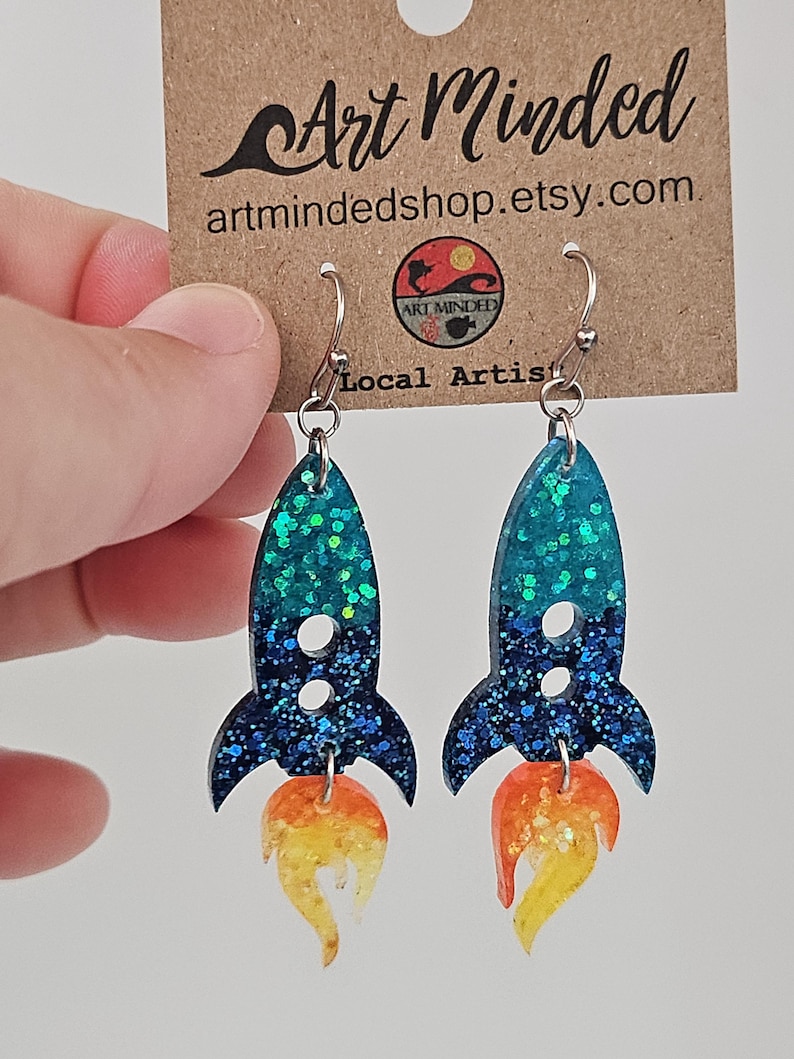 May include: A pair of handmade rocket ship earrings. The earrings feature a teal and blue glitter design with orange and yellow flames. The earrings are hanging from silver hooks. The background includes a brown tag with the text "Art Minded" and "artmindedshop.etsy.com."