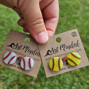 May include: Two pairs of glitter softball earrings. One pair is white with red stitching and the other is yellow with red stitching. The earrings are on a brown card with the text "Art Minded" and a website address.