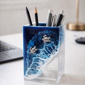 May include: A square desk organizer made of clear resin featuring a deep blue ocean wave design with white sea foam accents. Two small silver-toned sea turtle embellishments are attached to the front. The container holds several pens and pencils.