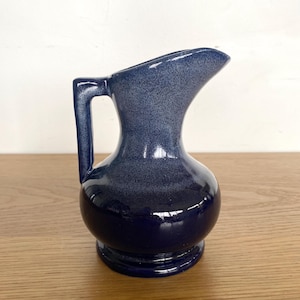 May include: A small, blue ceramic jug with a handle and spout. The jug has a speckled finish and a glossy, dark blue base. The handle is rectangular, and the spout is curved. The jug sits on a wooden surface.