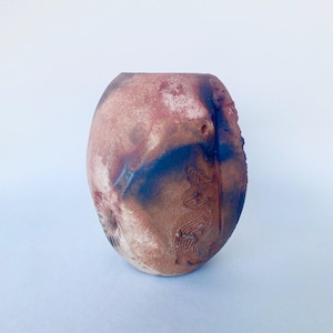 May include: A handcrafted ceramic vase with an oval form. The vase displays a mix of colours, including reddish-brown, blue, and white. The surface has textured details and a rough edge, enhancing its rustic appeal. The vase is suitable for home decor.