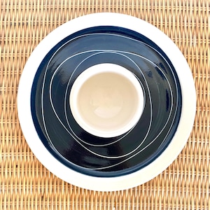 May include: A white ceramic plate with a black rim and a black centre with white swirling lines. A white ceramic bowl sits in the centre of the plate.