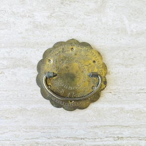 May include: Antique brass drawer pull with a floral, scalloped edge and a curved handle. The pull features an etched design of a dragon and other oriental motifs. The brass has a weathered, aged appearance.