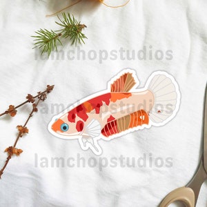May include: A sticker of a red, orange, and white betta fish with a white background. The sticker is on a white fabric surface with a spool of thread and a pair of scissors.
