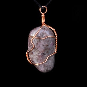 May include: A purple and gray stone pendant with a copper wire cage. The pendant is hanging from a black cord.