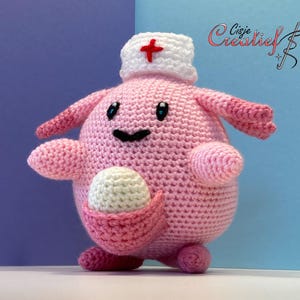 Op de afbeelding: Een handgemaakt roze gehaakt pluche speelgoed met een witte verpleegstershoed met een rood kruis. Het speelgoed heeft een lachend gezicht, kleine armen en een zakje met een wit eivormig voorwerp. De achtergrond is een verloop van blauw en paars.