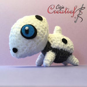 Op de afbeelding: Wit en grijs gehaakt amigurumi speelgoed met zwarte ogen en een blauw ooglapje. Het speelgoed heeft de vorm van een klein dier met een rond hoofd en een lange staart.