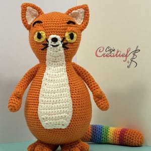 Op de afbeelding: Een gehaakt oranje vossenspeeltje met een crèmekleurige borst, gele ogen en zwarte geborduurde snorharen. De vos heeft puntige oren met crèmekleurige centra en een lange, gestreepte staart in regenboogkleuren. De tekst Cisje Creatief is zichtbaar.