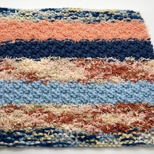 May include: A knitted dishcloth with a striped pattern in shades of blue, peach, and cream. The cloth has a textured, fluffy surface.