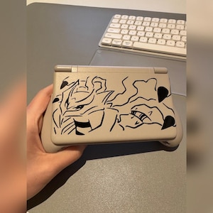 Puede incluir: Consola de juegos portátil beige con un diseño gráfico negro. La consola está abierta, mostrando la pantalla y los controles. Un teclado blanco es visible en el fondo.