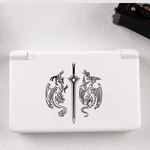 Puede incluir: Consola de juegos portátil blanca con un diseño de dragón y espada en blanco y negro. El diseño presenta dos dragones que flanquean una espada. La consola está cerrada y tiene un acabado liso y brillante.