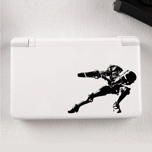 Può includere: Una console Nintendo DS Lite bianca con un adesivo in vinile in bianco e nero di un soldato futuristico in posa da combattimento. L'adesivo è sul lato inferiore destro della console.
