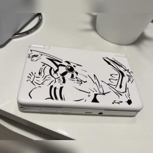 Puede incluir: Consola de juegos portátil blanca con un diseño gráfico negro. El diseño presenta una ilustración abstracta de una criatura. La consola está cerrada y apoyada sobre una superficie blanca.