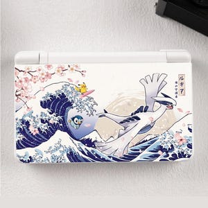 Pode incluir: Uma consola de jogos portátil branca com uma impressão gráfica de uma grande criatura branca a voar sobre ondas oceânicas azul-escuras estilizadas, uma personagem amarela numa prancha de surf, uma pequena personagem azul e flores de cerejeira.