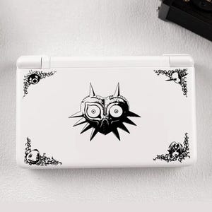 Puede incluir: Consola Nintendo DS Lite blanca con un diseño en blanco y negro de la Máscara de Majora. El diseño incluye la máscara en el centro y adornos decorativos en las esquinas. Una cámara negra es visible en el fondo.