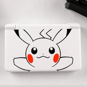 Puede incluir: Consola Nintendo DS Lite blanca con diseño de Pikachu. El diseño incluye un contorno negro de la cara, orejas, brazos y un rayo de Pikachu, con círculos rojos en las mejillas. La consola está cerrada y colocada sobre una superficie blanca.