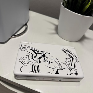May include: A white handheld gaming console featuring a black line art graphic of the legendary Pokémon Dialga on its top cover. The device is closed, showcasing the sleek white casing and the detailed black illustration of the creature.