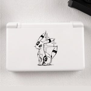 May include: White Nintendo DS Lite console with a black and white Umbreon illustration. The artwork is a detailed sketch. The console is closed and rests on a white surface. A black camera is visible in the background.