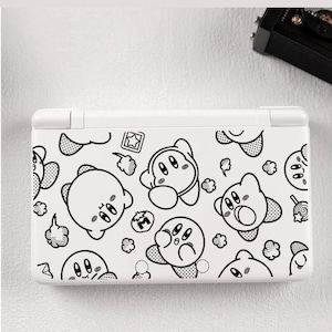 Puede incluir: Consola de juegos portátil blanca con un patrón repetido de personajes de dibujos animados en blanco y negro. El diseño incluye varias expresiones y elementos, creando una estética lúdica. La consola está cerrada y parece ser una Nintendo DS.
