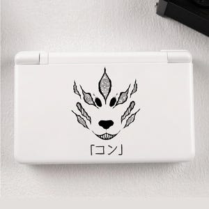 May include: White handheld gaming console featuring a black illustration of a stylized fox face with the Japanese characters 「コン」. The fox design includes intricate details and a smiling mouth. The console is closed and has a smooth, clean surface.