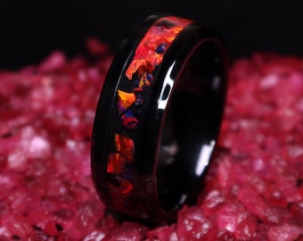 May include: A black ceramic ring with a band of red, orange, and blue opal inlay. The ring is photographed on a bed of red gemstones.