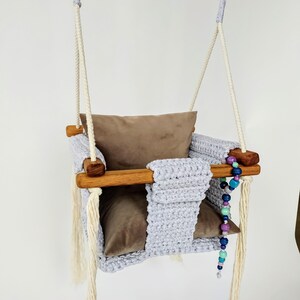 Könnte beinhalten: Ein grauer gehäkelter Babyschaukel mit einem braunen Samtkissen und einer Holzstange. Die Schaukel hat weiße Quasten und eine bunte Perlenkette, die daran hängt.
