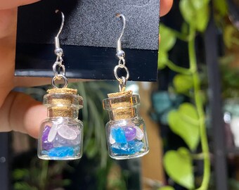 May include: A pair of silver dangle earrings with small glass bottles filled with colorful crystals. The bottles have cork stoppers and are suspended from silver hooks.