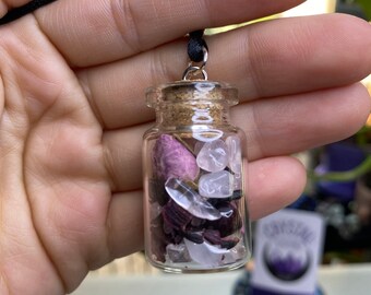 Könnte beinhalten: Eine kleine Glasflaschen-Halskette mit einem Korkverschluss. Die Flasche ist gefüllt mit rosa und weißen Kristallen, schwarzen Steinen und getrockneten rosa Blüten.