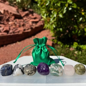 May include: Set of seven tumbled stones, including blue, white, gold, purple, green, pink, and green, arranged on a white surface. The stones are in a green velvet pouch.