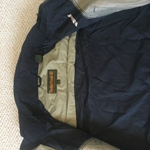 Könnte beinhalten: Eine marineblaue und beige Jacke mit Reißverschluss. Die Jacke hat ein Mesh-Futter und ein Etikett mit der Aufschrift "Timberland Weathergear".