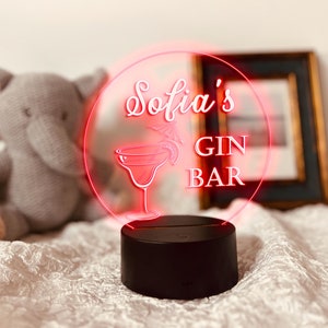K&ouml;nnte beinhalten: Ein schwarzer Sockel tr&auml;gt ein rundes, beleuchtetes Schild mit den Worten "Sofia's GIN BAR" in wei&szlig;er Schrift und einer Margarita-Glas-Illustration. Das Schild leuchtet mit einem roten Umriss. Ein Stoffelefant-Spielzeug befindet sich im Hintergrund.