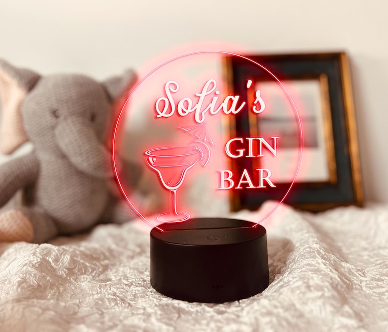 K&ouml;nnte beinhalten: Ein schwarzer Sockel tr&auml;gt ein rundes, beleuchtetes Schild mit den Worten "Sofia's GIN BAR" in wei&szlig;er Schrift und einer Margarita-Glas-Illustration. Das Schild leuchtet mit einem roten Umriss. Ein Stoffelefant-Spielzeug befindet sich im Hintergrund.
