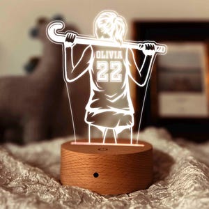 Puede incluir: Una base de madera con una silueta blanca de una persona sosteniendo un palo de hockey. La silueta lleva una camiseta con el nombre "Olivia" y el número "22" en la espalda.