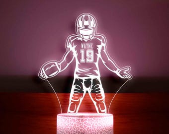 Pode incluir: Uma luz noturna impressa em 3D branca em forma de jogador de futebol americano vestindo um capacete e uma camisa com o número 19 e o nome "WAYNE" nela. A luz noturna está sobre uma base branca com uma superfície texturizada.