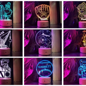 May include: Nine different personalized night lights with various designs, including a baseball glove, a sleeping bear, a construction crane, a video game, a popcorn bucket, a saxophone, and a Santa Claus. Each light has a wooden base and a clear acrylic panel with a white design.