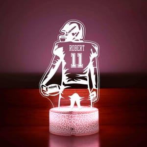 Puede incluir: Silueta blanca de un jugador de fútbol americano sosteniendo un balón de fútbol con el nombre "ROBERT" y el número "11" en la camiseta. La silueta está sobre una base blanca con una superficie texturizada. La base está iluminada desde el interior con una luz blanca cálida.