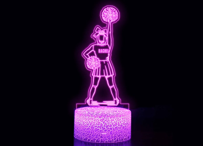 May include: A pink 3D printed night light shaped like a cheerleader holding a pom-pom. The cheerleader is wearing a skirt and has the name "Rachel" on her shirt.