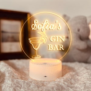 K&ouml;nnte beinhalten: Ein leuchtendes LED-Schild mit der Aufschrift "Sofia's GIN BAR" in einem kreisf&ouml;rmigen Design. Das Schild zeigt eine Cocktailglas-Illustration und einen dekorativen Schirm. Der wei&szlig;e Sockel hat eine Rissstruktur.