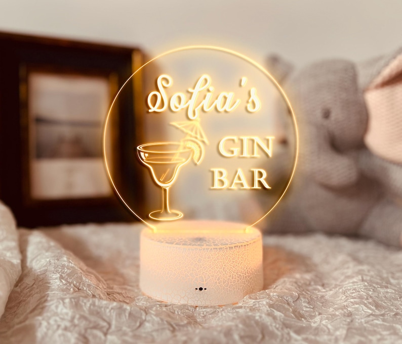 K&ouml;nnte beinhalten: Ein leuchtendes LED-Schild mit der Aufschrift "Sofia's GIN BAR" in einem kreisf&ouml;rmigen Design. Das Schild zeigt eine Cocktailglas-Illustration und einen dekorativen Schirm. Der wei&szlig;e Sockel hat eine Rissstruktur.