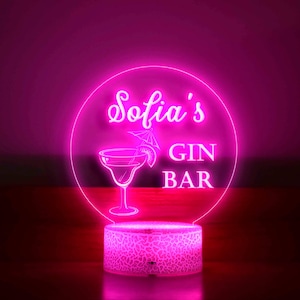 K&ouml;nnte beinhalten: Ein neonrosa LED-Schild mit der Aufschrift "Sofia's GIN BAR" in einem kreisf&ouml;rmigen Design. Das Schild zeigt eine Cocktailglas-Illustration und steht auf einem gemusterten Sockel, der vor einem dunklen Hintergrund leuchtet.
