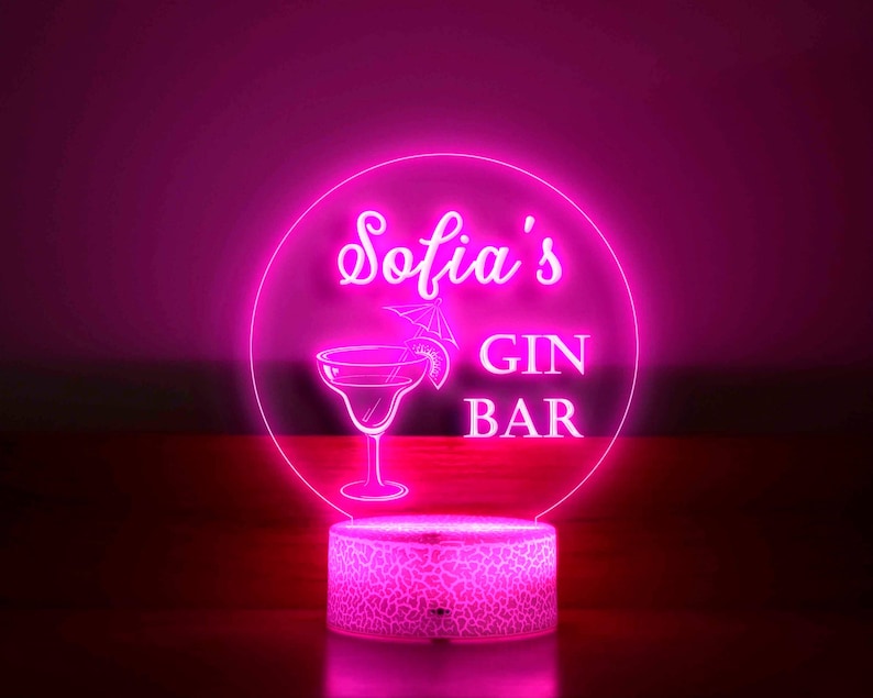 K&ouml;nnte beinhalten: Ein neonrosa LED-Schild mit der Aufschrift "Sofia's GIN BAR" in einem kreisf&ouml;rmigen Design. Das Schild zeigt eine Cocktailglas-Illustration und steht auf einem gemusterten Sockel, der vor einem dunklen Hintergrund leuchtet.