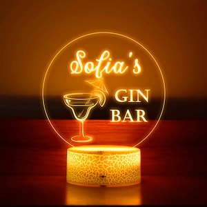 May include: A glowing LED sign with the text "Sofia's GIN BAR" in a circular design. The sign features a margarita glass illustration and a decorative base with a textured pattern. The sign emits a warm, orange light.
