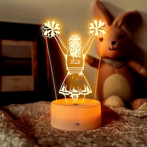 Könnte beinhalten: Eine leuchtende LED-Nachtlampe mit einem Cheerleader-Design. Das Licht zeigt eine Cheerleader-Figur mit Pompons, und der Name "JULIA" ist auf der Uniform zu sehen. Der Sockel ist ein strukturierter, orangefarbener Zylinder.
