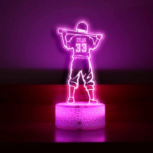 May include: A pink 3D printed night light shaped like a baseball player holding a bat. The player is wearing a jersey with the name "DYLAN" and the number "33" on the back. The night light has a textured base.