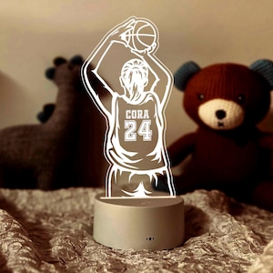 Könnte beinhalten: Eine weiße LED-Lampe mit einer Basketballspieler-Silhouette, dem Namen "CORA" und der Nummer "24" auf dem Trikot. Die Lampe steht auf einem hellgrauen Sockel. Ein brauner Teddybär und ein graues Dinosaurier-Plüschtier sind im Hintergrund.