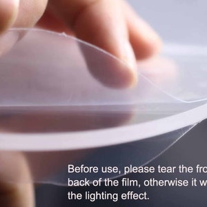 May include: A hand peeling off a clear protective film from a round object. The text on the image reads: "Before use, please tear the front and back of the film, otherwise it will affect the lighting effect."