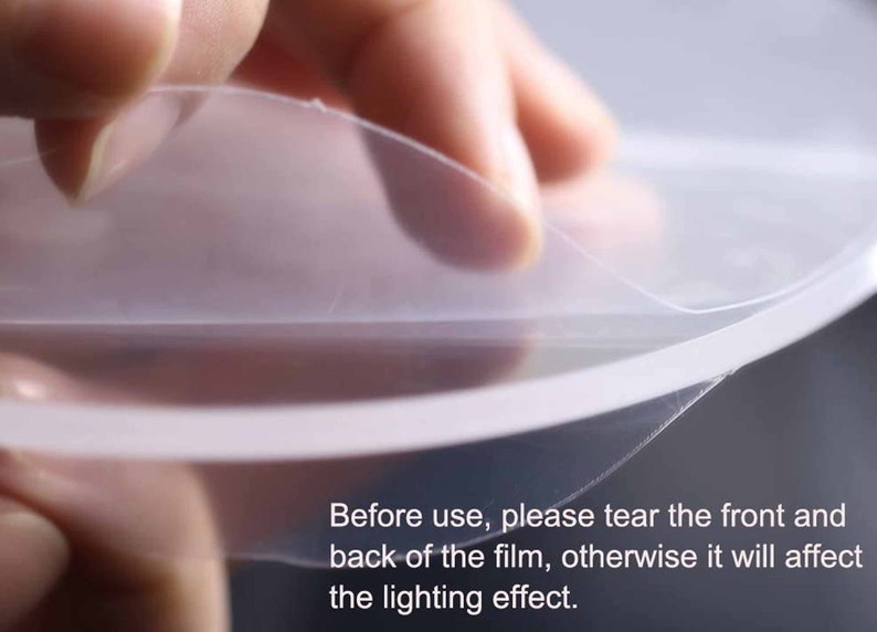 May include: A hand peeling off a clear protective film from a round object. The text on the image reads: "Before use, please tear the front and back of the film, otherwise it will affect the lighting effect."