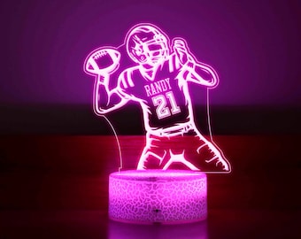 Pode incluir: Uma luz noturna LED rosa em forma de jogador de futebol americano lançando uma bola de futebol. O jogador está vestindo uma camisa com o nome "RANDY" e o número "21".