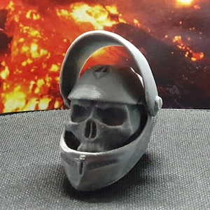 May include: A gray 3D printed skull wearing a helmet with a visor. The helmet has a raised design on the top.
