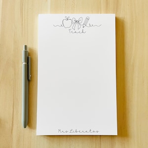 Op de afbeelding: Een witte notitieblok met een zwarte lijntekening van een appel, strik en potlood, met het woord "Teach" en de naam "Mrs. Liberatos" eronder gedrukt. Een grijze pen ligt links van het notitieblok.